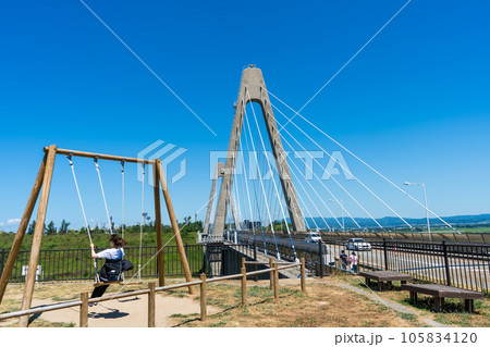 晴れの夏空と日本海と河北潟を繋ぐ河北潟放水路に架かる内灘大橋と道の駅のブランコ｜石川県河北郡内灘町 105834120