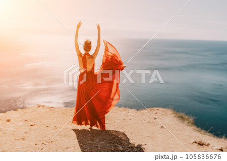 Woman in red dress on sea. Side view a Young beautiful sensual woman in a red long dress posing on a rock high above the sea on sunset. Girl on the nature on blue sky background. Fashion photo. Woman in red dress on sea. Side view a Young beautiful sensual woman in a red long dress posing on a rock high above the sea on sunset. Girl on the nature on blue sky background. Fashion photo. 105836676