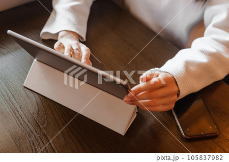 A girl at a wooden table in a restaurant is working on a tablet. Women's hands close-up. A girl at a wooden table in a restaurant is working on a tablet. Women's hands close-up. 105837982