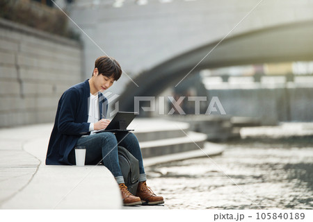 A young man working on a laptop in a park with a river A young man working on a laptop in a park with a river 105840189