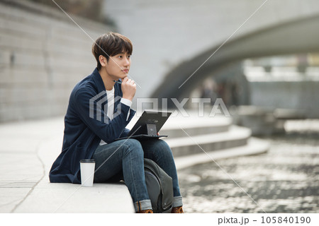 A young man working on a laptop in a park with a river A young man working on a laptop in a park with a river 105840190