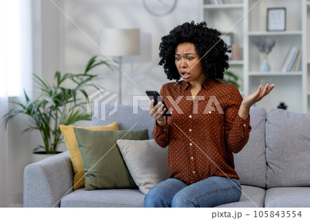 Sad woman alone at home, african american woman received notification message with bad news sitting on couch in living room, holding phone in hands using social media app. Sad woman alone at home, african american woman received notification message with bad news sitting on couch in living room, holding phone in hands using social media app. 105843554