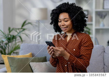 Young beautiful African American woman sitting on sofa at home, woman with curly hair holding phone in hands, using online application in living room browsing internet pages and typing messages 105843567