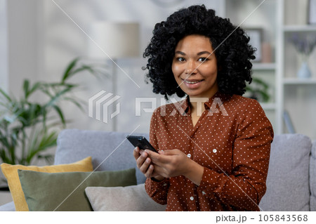 Portrait of a young successful woman at home, an African American woman smiling and looking at the camera, holding the phone in her hands while sitting on the couch in the living room. Portrait of a young successful woman at home, an African American woman smiling and looking at the camera, holding the phone in her hands while sitting on the couch in the living room. 105843568