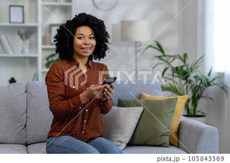 Portrait of a young successful woman at home, an African American woman smiling and looking at the camera, holding the phone in her hands while sitting on the couch in the living room. 105843569