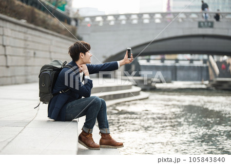 A young man relaxing by the river in the city and taking pictures with his smartphone 105843840