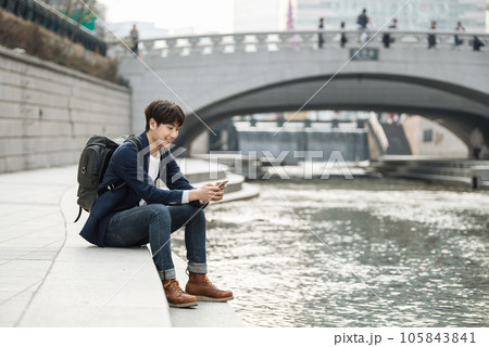 A young man relaxing by the river in the city and taking pictures with his smartphone 105843841