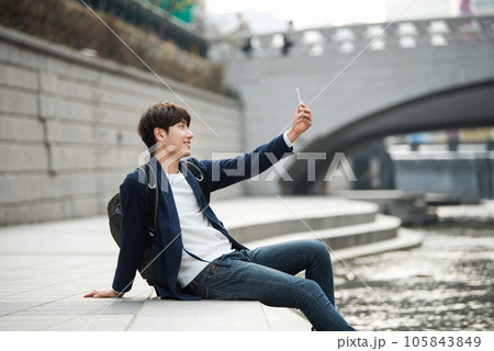 A young man relaxing by the river in the city and taking pictures with his smartphone 105843849