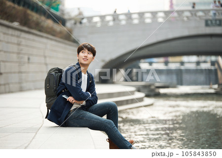 A young man relaxing by the river in the city and taking pictures with his smartphone 105843850