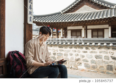A young man who uses a tablet while traveling in tourist attractions with traditional Korean houses A young man who uses a tablet while traveling in tourist attractions with traditional Korean houses 105845737