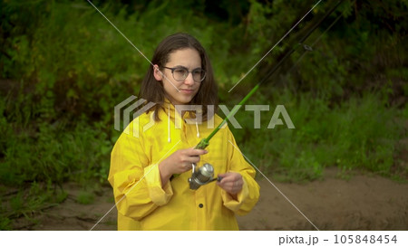 A young woman is fishing on the river bank. A girl in a yellow raincoat with a hood and glasses casts a spinning rod. Close-up. A young woman is fishing on the river bank. A girl in a yellow raincoat with a hood and glasses casts a spinning rod. Close-up. 105848454