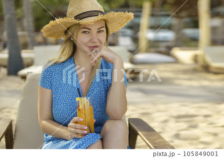 Fashion young woman drinking cocktail in beach bar. Cheerful attractive blonde woman in a dress smiles and drinks a cocktail outdoors. Happy woman enjoying summer time at the beach Fashion young woman drinking cocktail in beach bar. Cheerful attractive blonde woman in a dress smiles and drinks a cocktail outdoors. Happy woman enjoying summer time at the beach 105849001
