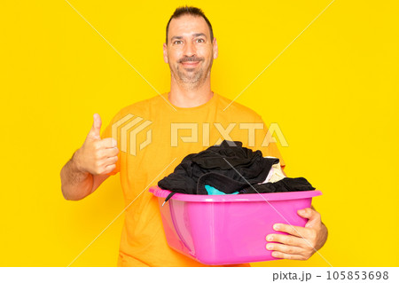 Bearded hispanic man doing housework holding plastic basket with clothes raising thumb up, excellent symbol. Isolated on yellow background. Bearded hispanic man doing housework holding plastic basket with clothes raising thumb up, excellent symbol. Isolated on yellow background. 105853698