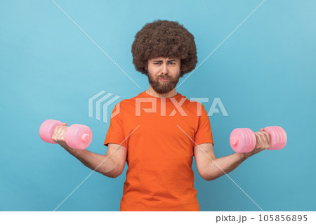 Portrait of funny man with Afro hairstyle wearing orange T-shirt rising hands holding dumbbells, pumping up muscles, having serious silly expression. Indoor studio shot isolated on blue background. 105856895