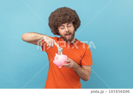 Portrait of positive happy man with Afro hairstyle wearing orange T-shirt investing, putting dollar banknote into piggy bank, money savings. Indoor studio shot isolated on blue background. 105856896
