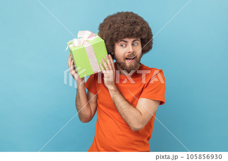 Portrait of curious man with Afro hairstyle wearing orange T-shirt standing with green wrapped present box and shacking gift, what inside. Indoor studio shot isolated on blue background. Portrait of curious man with Afro hairstyle wearing orange T-shirt standing with green wrapped present box and shacking gift, what inside. Indoor studio shot isolated on blue background. 105856930