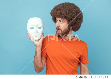 Portrait of man with Afro hairstyle wearing orange T-shirt looking at white mask in hands with attentive look, trying to understand hiding personality. Indoor studio shot isolated on blue background. Portrait of man with Afro hairstyle wearing orange T-shirt looking at white mask in hands with attentive look, trying to understand hiding personality. Indoor studio shot isolated on blue background. 105856931