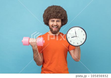 Portrait of man with Afro hairstyle wearing orange T-shirt holding dumbbell and big wall clock, it's time for sport, starting your trainings. Indoor studio shot isolated on blue background. Portrait of man with Afro hairstyle wearing orange T-shirt holding dumbbell and big wall clock, it's time for sport, starting your trainings. Indoor studio shot isolated on blue background. 105856932