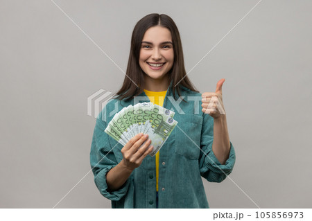 Smiling dark haired woman holding many euro banknotes and showing thumb up to camera, satisfied of winning lottery, wearing casual style jacket. Indoor studio shot isolated on gray background. 105856973