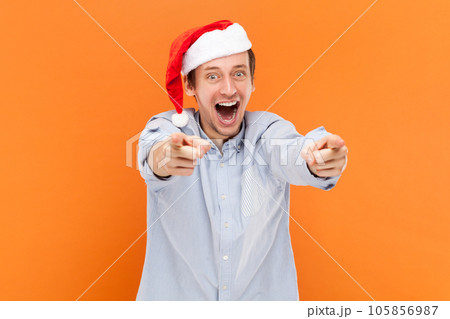 Portrait of amazed handsome young adult man standing pointing fingers to camera, choosing you, wearing light blue shirt and Santa Claus hat. Indoor studio shot isolated on orange background. 105856987