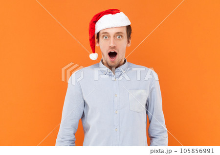 Shocked astonished young adult man looking at camera with widely open mouth, sees something surprised, wearing light blue shirt and Santa Claus hat. Indoor studio shot isolated on orange background. 105856991