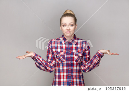 Portrait of uncertain confused attractive woman with bun hairstyle shrugging shoulders, dosen't know answer, wearing checkered shirt. Indoor studio shot isolated on gray background. Portrait of uncertain confused attractive woman with bun hairstyle shrugging shoulders, dosen't know answer, wearing checkered shirt. Indoor studio shot isolated on gray background. 105857016