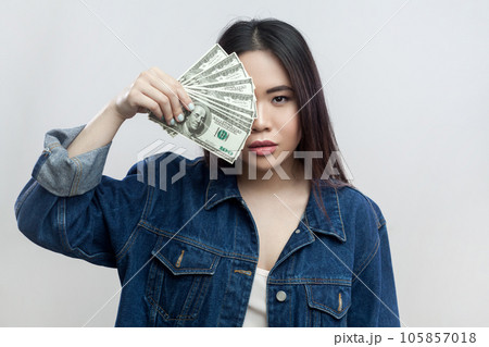 Portrait of beautiful brunette woman in blue denim jacket standing covering half of her hair with big fan of dollar banknotes, betting, winning. Indoor studio shot isolated on gray background. 105857018