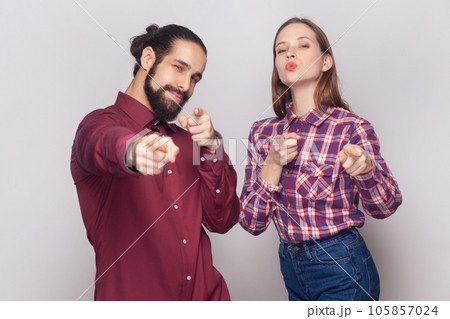 Portrait of satisfied delighted cute woman and man standing together pointing at camera, selecting you, expressing positive emotions. Indoor studio shot isolated on gray background. 105857024