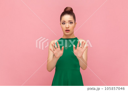 Portrait of scared frighten woman with bun hairstyle standing showing palms to camera, demonstrates stop sign, asking to stop it, wearing green dress. Indoor studio shot isolated on pink background. 105857046