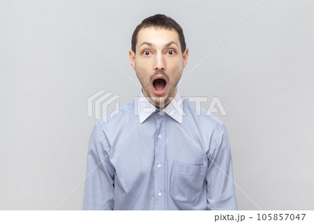 Portrait of shocked astonished surprised man looking at camera with big eyes and open mouth, sees something amazed, wearing light blue shirt. Indoor studio shot isolated on gray background. Portrait of shocked astonished surprised man looking at camera with big eyes and open mouth, sees something amazed, wearing light blue shirt. Indoor studio shot isolated on gray background. 105857047