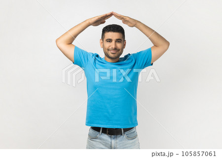 Portrait of positive friendly smiling unshaven man wearing blue T- shirt standing making roof gesture, feeling himself in safety. Indoor studio shot isolated on gray background. 105857061