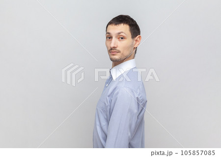 Side view portrait of serious attractive man standing looking at camera with strict concentrated facial expression, wearing light blue shirt. Indoor studio shot isolated on gray background. 105857085