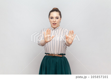 Portrait of scared frighten young adult woman wearing striped shirt and green skirt sees something scary, showing stop palm gesture. Indoor studio shot isolated on gray background. 105857101