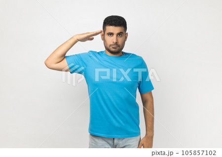 Portrait of serious strict handsome unshaven man wearing blue T- shirt standing with hand near, saying yes sir, being ready to do command. Indoor studio shot isolated on gray background. Portrait of serious strict handsome unshaven man wearing blue T- shirt standing with hand near, saying yes sir, being ready to do command. Indoor studio shot isolated on gray background. 105857102