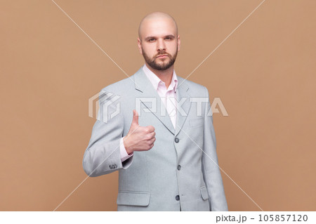 Portrait of serious attractive bald bearded man standing showing thumb up, recommend good service, positive feedback, wearing gray jacket. Indoor studio shot isolated on brown background. Portrait of serious attractive bald bearded man standing showing thumb up, recommend good service, positive feedback, wearing gray jacket. Indoor studio shot isolated on brown background. 105857120