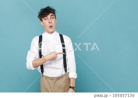 Portrait of amazed surprised man wearing white shirt and suspender, pointing aside with finger, showing copy space for promotion. Indoor studio shot isolated on blue background. Portrait of amazed surprised man wearing white shirt and suspender, pointing aside with finger, showing copy space for promotion. Indoor studio shot isolated on blue background. 105857130