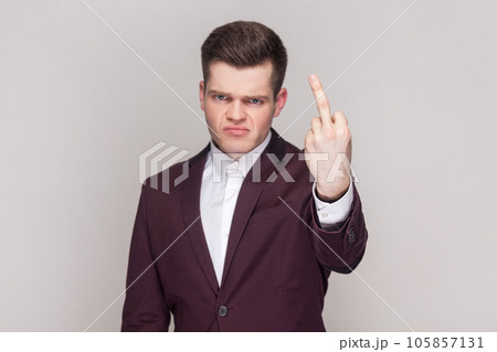 Portrait of man shows fuck off sign being rude and looks dissatisfied at camera, expresses bad attitude to someone, wearing violet suit and white shirt. Indoor studio shot isolated on grey background. 105857131