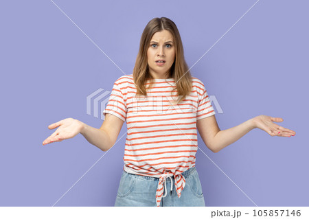 I don't know. Blond woman wearing striped T-shirt standing with wide raised arms and looking at camera with frowning face, and dont know what to do. Indoor studio shot isolated on purple background. I don't know. Blond woman wearing striped T-shirt standing with wide raised arms and looking at camera with frowning face, and dont know what to do. Indoor studio shot isolated on purple background. 105857146