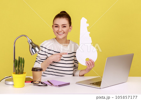 Amazing woman sitting at workplace with laptop, pointing at big paper key and smiling, satisfied with new home, rental office. Indoor studio studio shot isolated on yellow background. 105857177