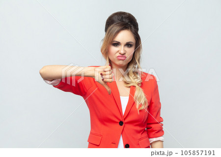 Portrait of disappointed attractive upset sad blonde woman standing showing thumb down, dislike gesture, disagree, wearing red jacket. Indoor studio shot isolated on gray background. 105857191