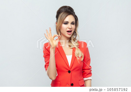 Portrait of playful cheerful beautiful young adult blonde woman standing showing ok sign, making okay gesture with hand, wearing red jacket. Indoor studio shot isolated on gray background. 105857192