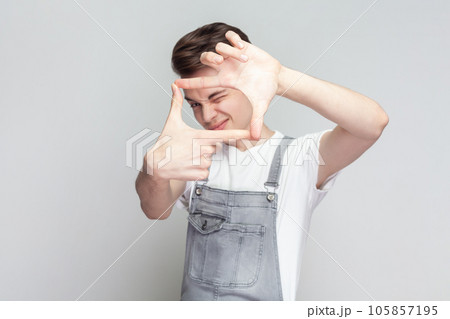Gesturing finger frame. Portrait of young brunette man standing looking at camera and gesturing finger frame, wearing denim overalls. Indoor studio shot isolated on gray background. Gesturing finger frame. Portrait of young brunette man standing looking at camera and gesturing finger frame, wearing denim overalls. Indoor studio shot isolated on gray background. 105857195
