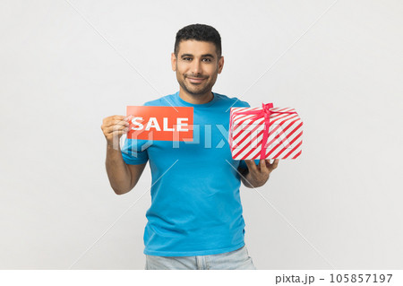 Portrait of positive optimistic unshaven man wearing blue T- shirt standing with present gift box and sale card, discounts for presents before holidays. Indoor studio shot isolated on gray background. 105857197