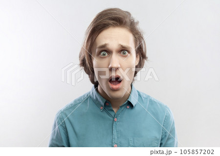Portrait of shocked amazed astonished young man looking at camera with big eyes and open mouth, sees something amazing, wearing blue shirt. Indoor studio shot isolated on gray background. 105857202