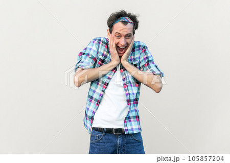 Wow, that great. Overjoyed handsome man keeps hands on cheeks, looks joyfully at camera, being amazed, wearing blue checkered shirt and headband. Indoor studio shot isolated on gray background. 105857204