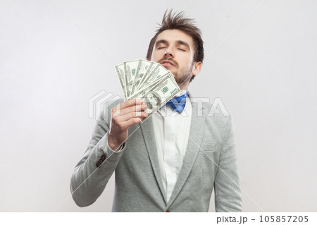 Portrait of rich delighted proud bearded man standing smelling dollar banknotes, being satisfied of big salary, wearing grey suit and blue bow tie. Indoor studio shot isolated on gray background. Portrait of rich delighted proud bearded man standing smelling dollar banknotes, being satisfied of big salary, wearing grey suit and blue bow tie. Indoor studio shot isolated on gray background. 105857205