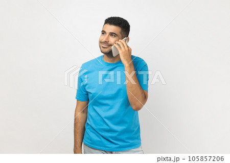 Portrait of optimistic smiling unshaven man in blue T- shirt standing talking on cell phone, having pleased positive expression, enjoying conversation. Indoor studio shot isolated on gray background. Portrait of optimistic smiling unshaven man in blue T- shirt standing talking on cell phone, having pleased positive expression, enjoying conversation. Indoor studio shot isolated on gray background. 105857206