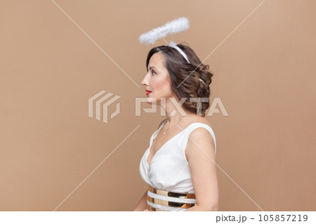 Side view portrait of calm attractive middle aged woman with wavy hair and nimb over head, looking ahead, wearing white dress. Indoor studio shot isolated on light brown background. Side view portrait of calm attractive middle aged woman with wavy hair and nimb over head, looking ahead, wearing white dress. Indoor studio shot isolated on light brown background. 105857219