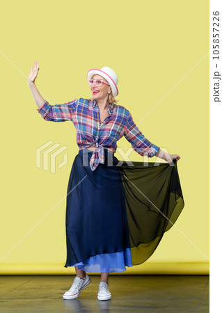 Full length portrait of mature woman wearing hat and eyeglasses standing, posing, looking away and waving hand, greeting somebody. Indoor studio shot isolated on yellow background Full length portrait of mature woman wearing hat and eyeglasses standing, posing, looking away and waving hand, greeting somebody. Indoor studio shot isolated on yellow background 105857226