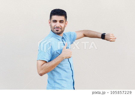 Portrait of positive attractive calm man wearing denim shirt standing showing his smart watch and thumb up, like gesture, recommend. Indoor studio shot isolated on gray background. Portrait of positive attractive calm man wearing denim shirt standing showing his smart watch and thumb up, like gesture, recommend. Indoor studio shot isolated on gray background. 105857229
